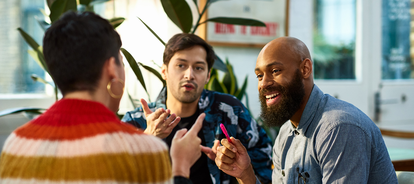 Three people in casual discussion indoors with plants and framed art in background